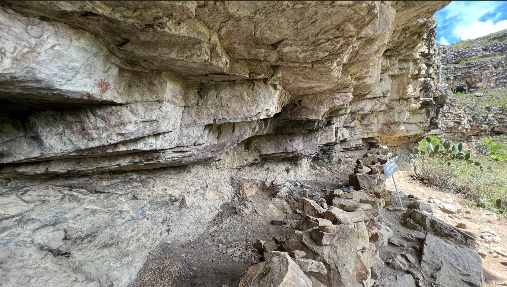 The Sáchica rock shelter — red pictograms visible on the cliff face beneath the protective overhang, with the Boyacá landscape stretching beyond